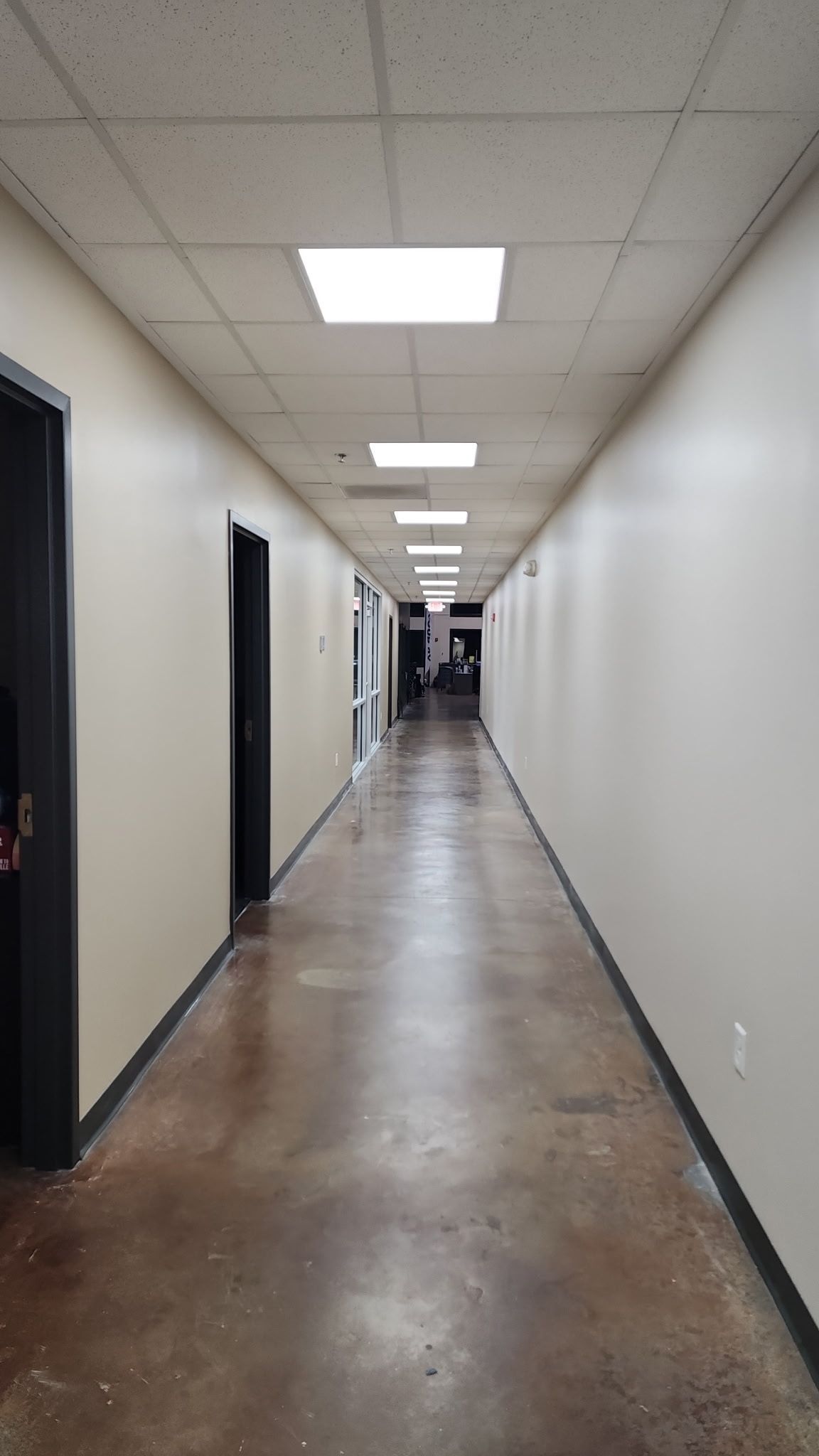 Long office hallway with beige walls, dark doorways, and fluorescent ceiling lights.