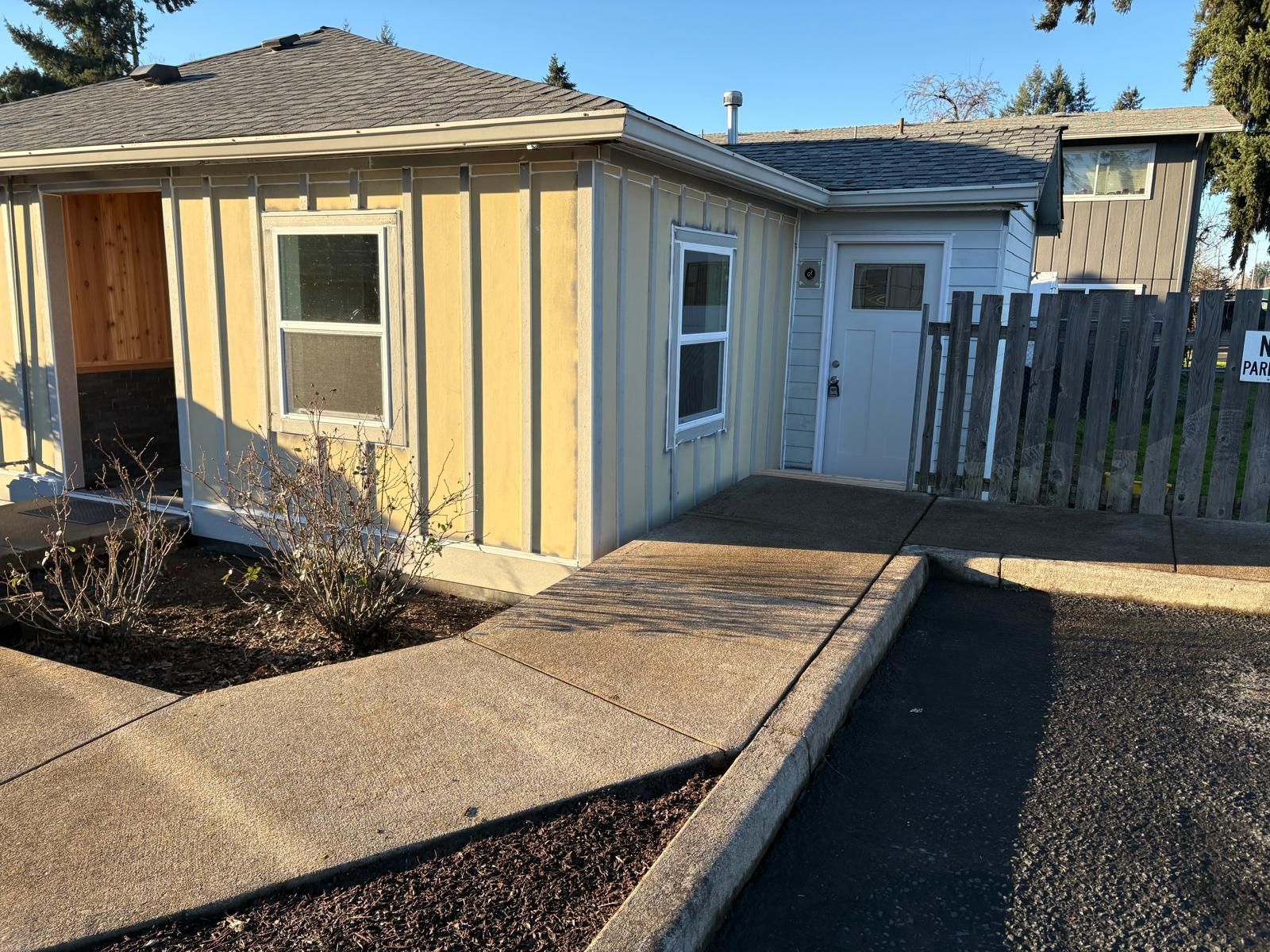 Small yellow house with a white door, window, fenced side yard, and concrete walkway in daylight