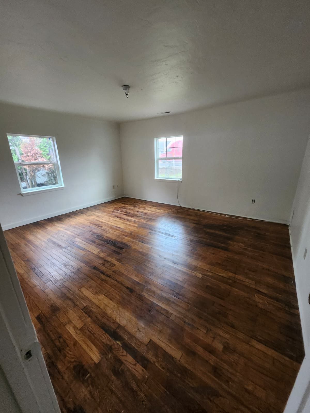 Empty room with white walls, hardwood floors, and two windows letting in daylight.