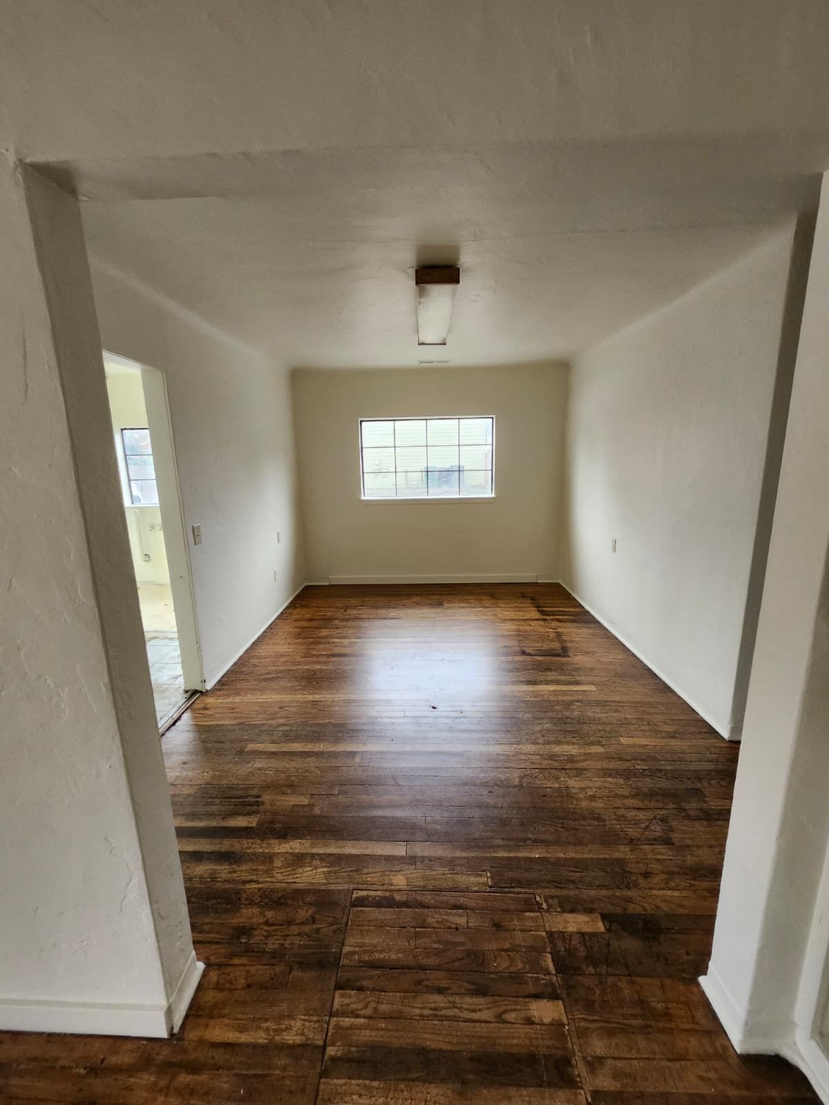 Empty narrow room with white walls, dark wood floor, ceiling light, and a small window at the far end.