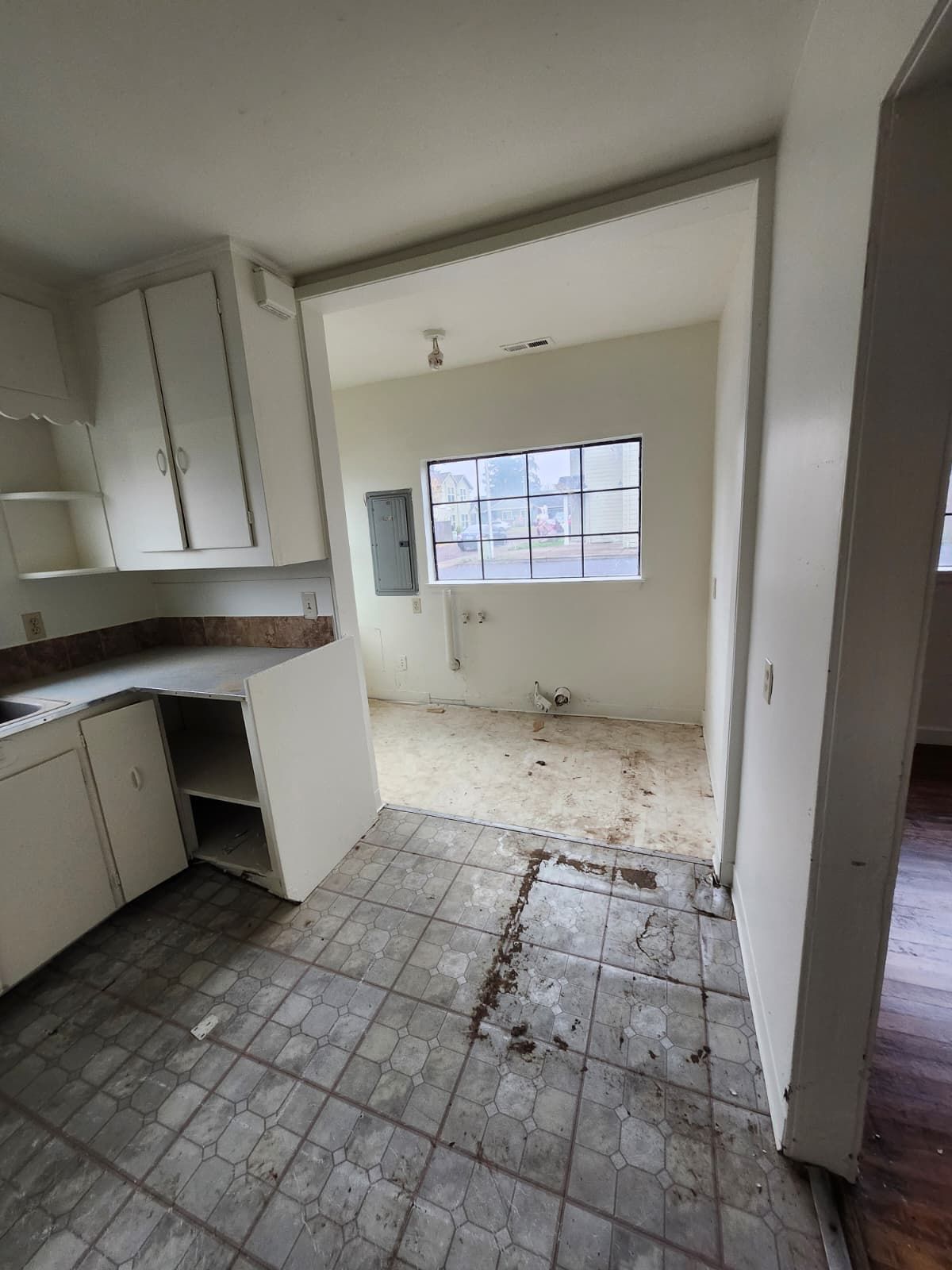 Empty, damaged kitchen with white cabinets, tiled floor, and debris near a window.