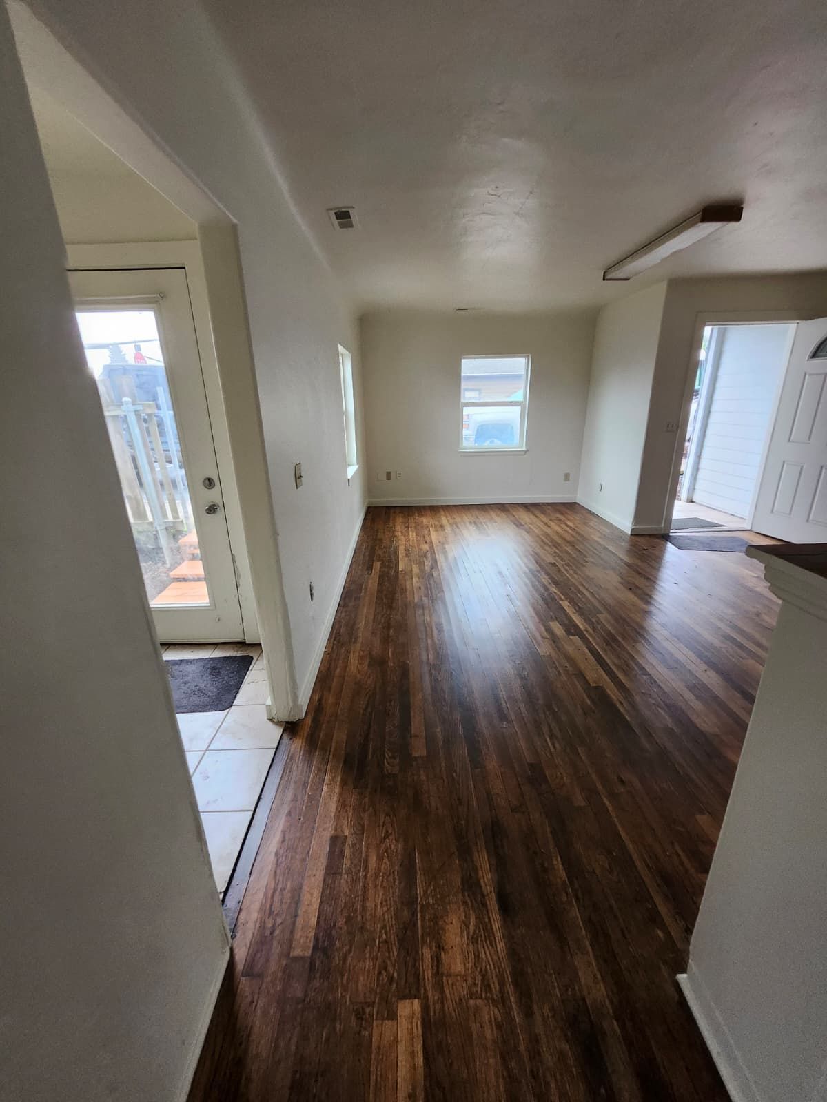 Empty apartment room with dark wood floors, white walls, and a window letting in daylight