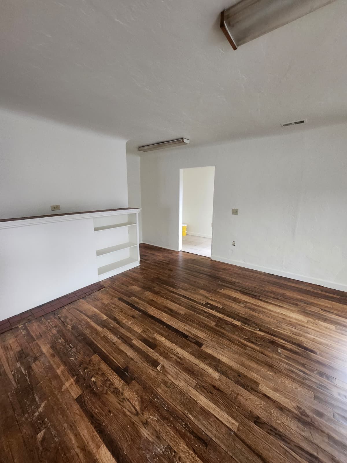 Empty room with dark wood floors, white walls, and a doorway leading to another bright room