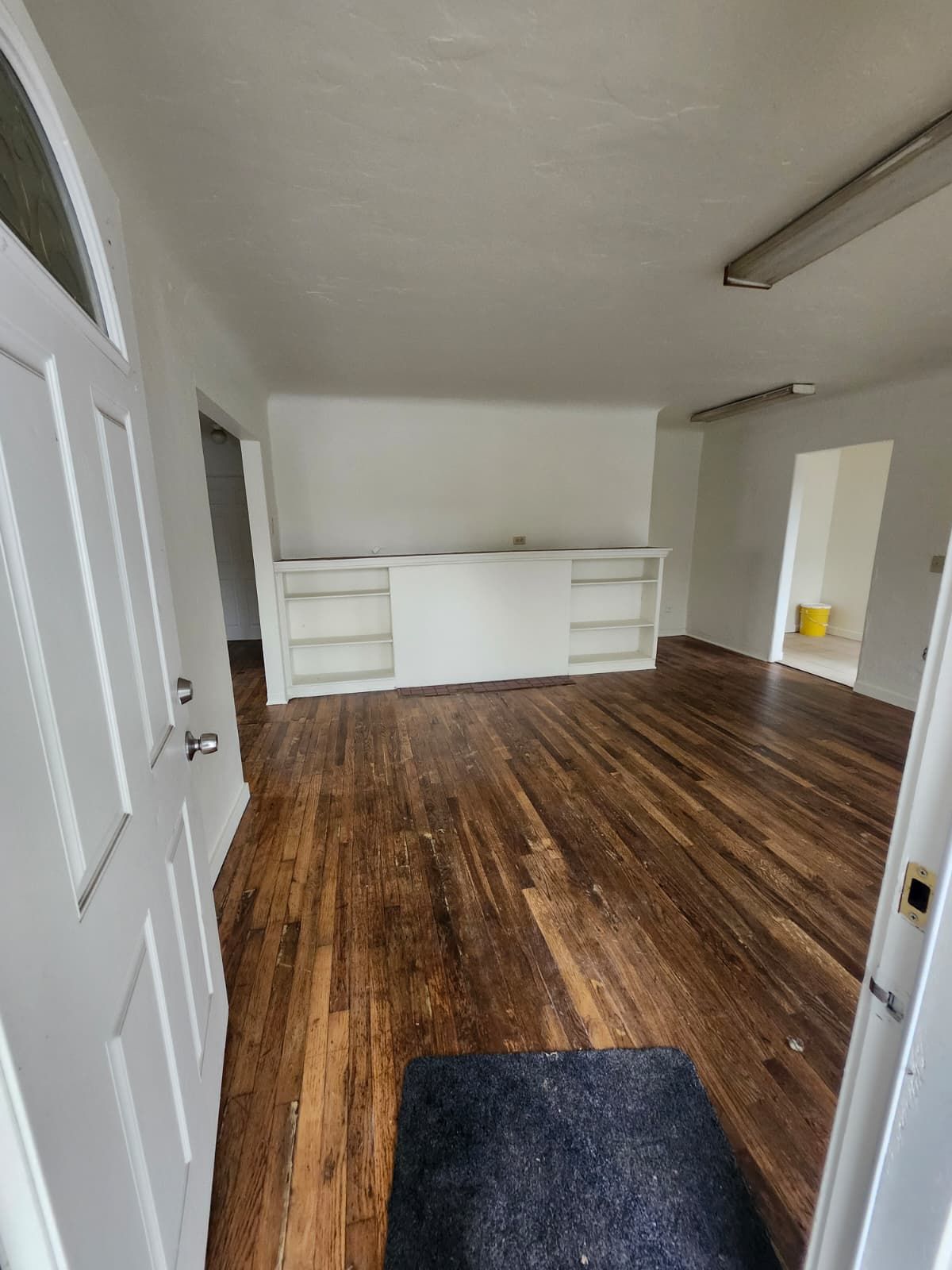 Empty room with white walls, dark wood floor, built-in shelves, and a doorway to a yellow-lit space