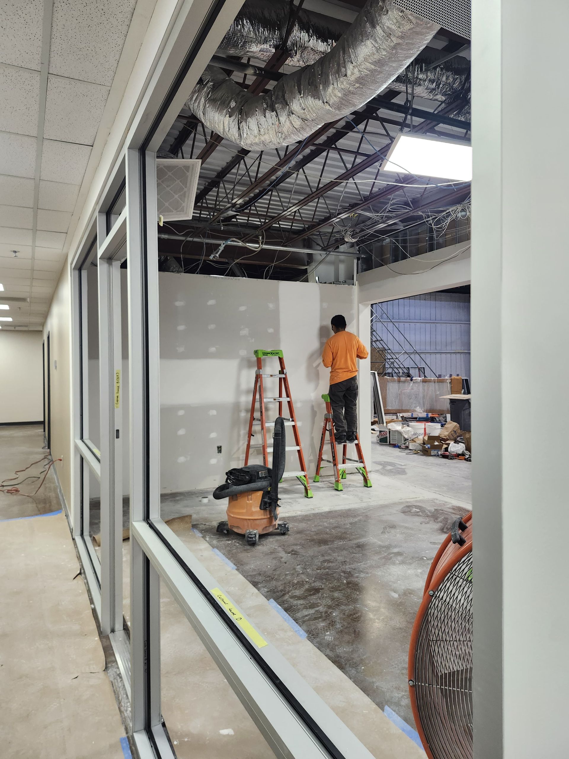 Construction worker on ladder in unfinished office with exposed ceiling and drywall.
