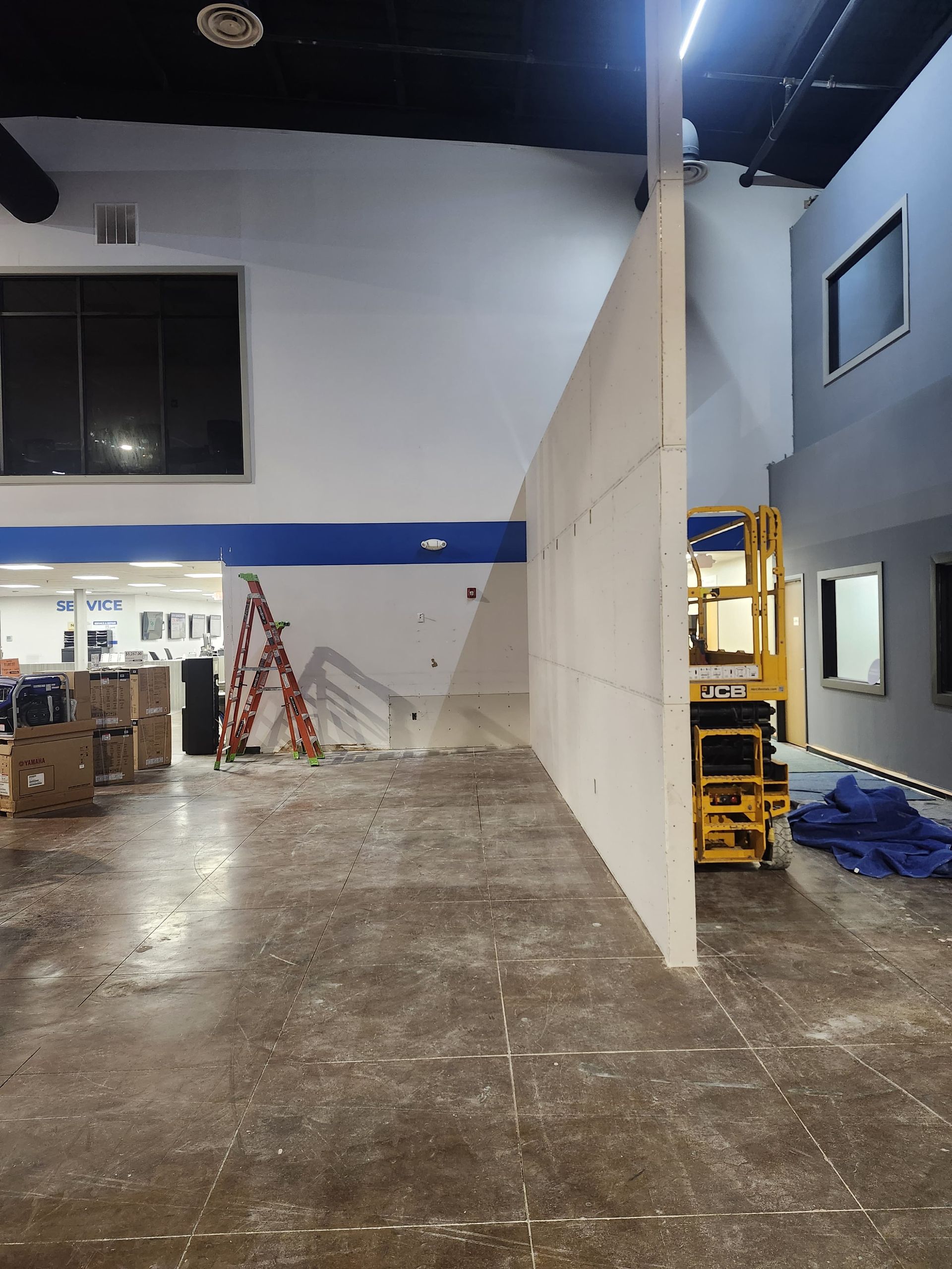 Interior of a warehouse-like room with wet concrete floor, a white partition wall, ladder, and yellow forklift.
