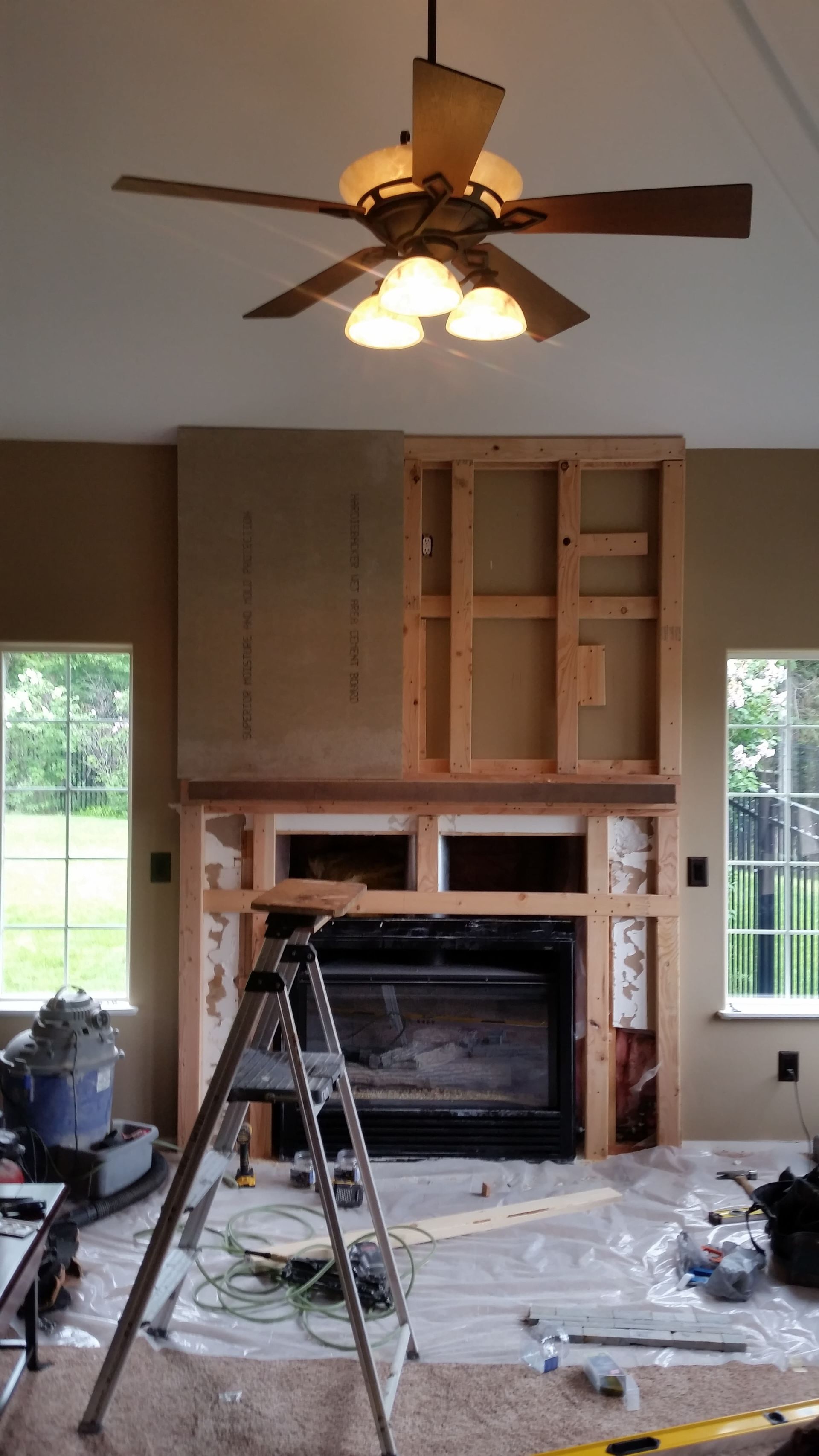Living room under renovation with built-in shelves, fireplace, ladder, debris, and ceiling fan light on