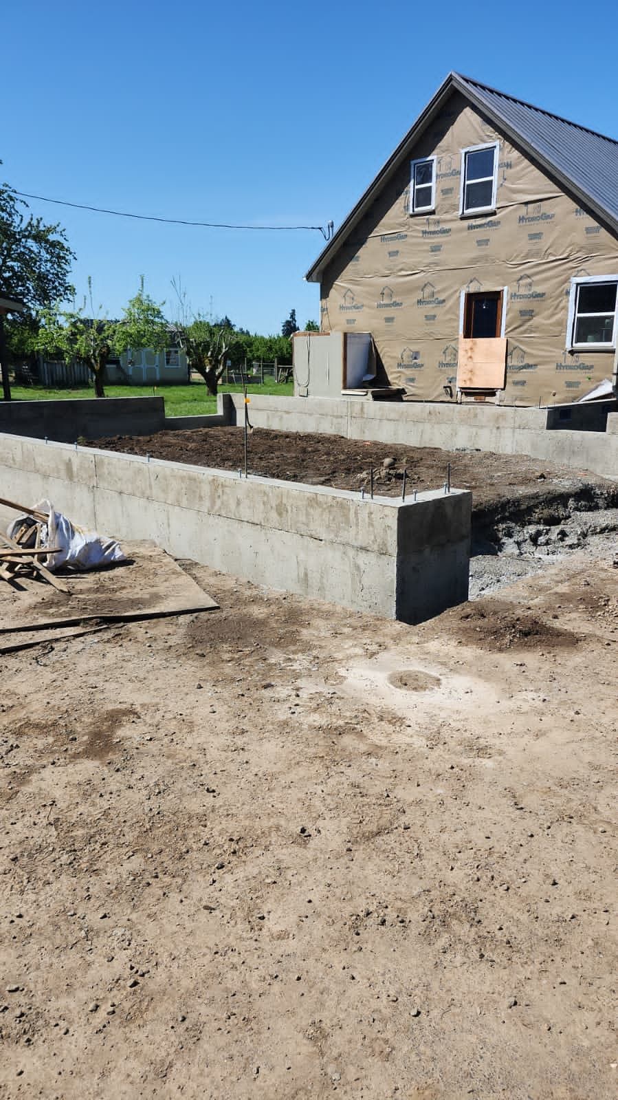 Stone house under construction with a concrete foundation and dirt foreground, viewed on a sunny day