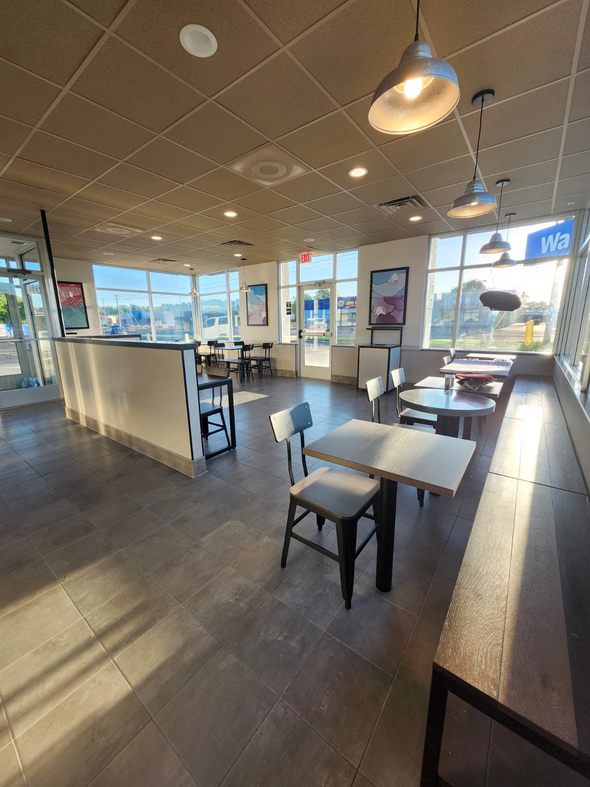 Empty fast-food dining area with white tables, gray chairs, and large windows on a tiled floor.