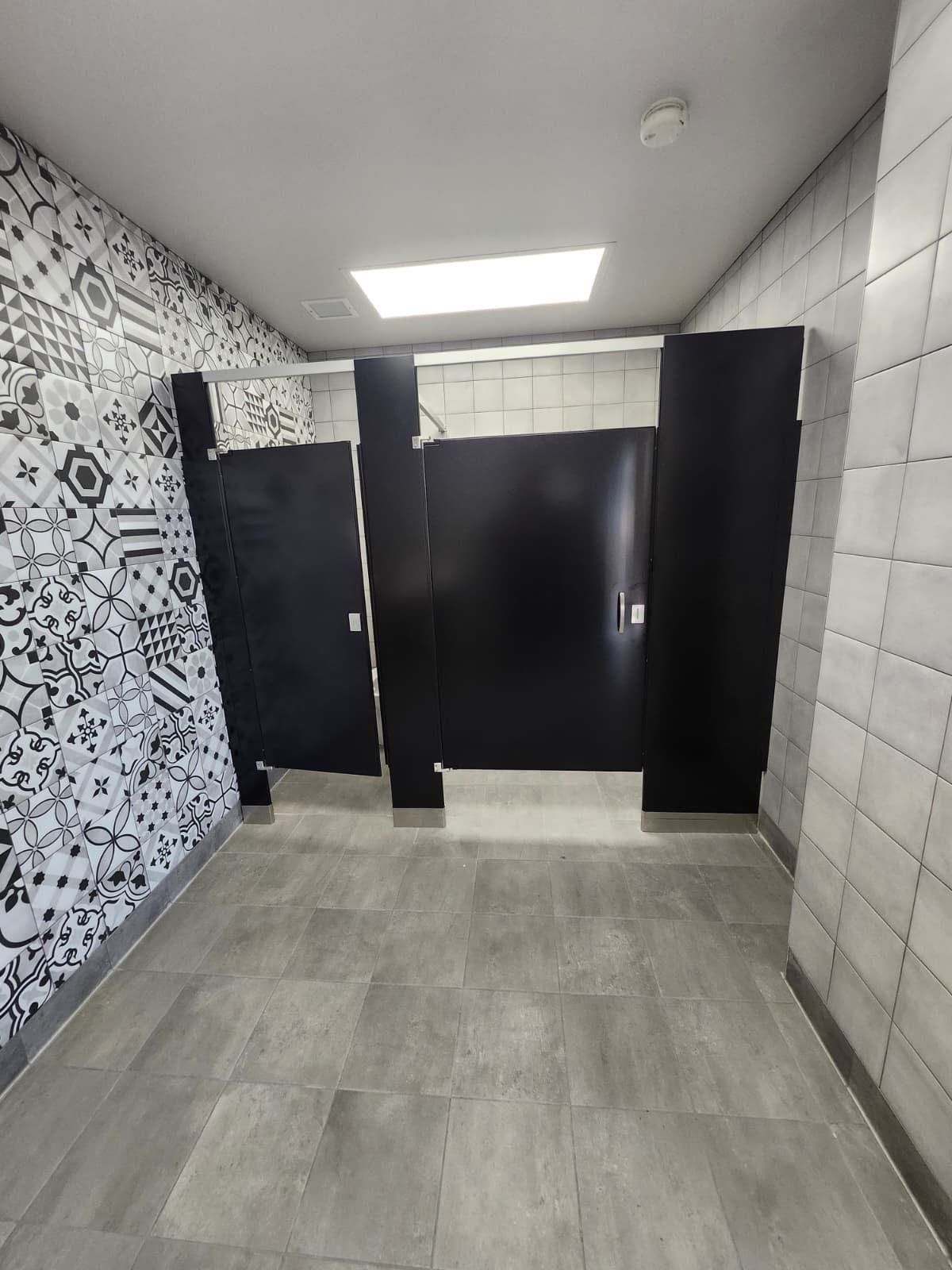 Public restroom with tiled walls, gray floor, black stall doors, and a bright ceiling light