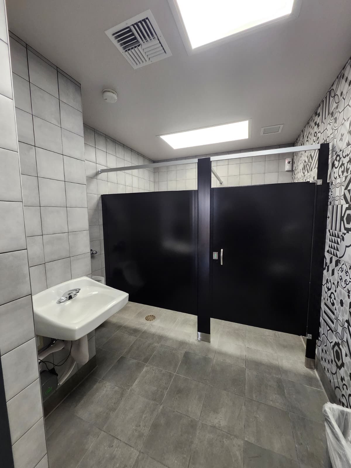 Small restroom with black stalls, wall-mounted sink, gray tile, and ceiling lights.