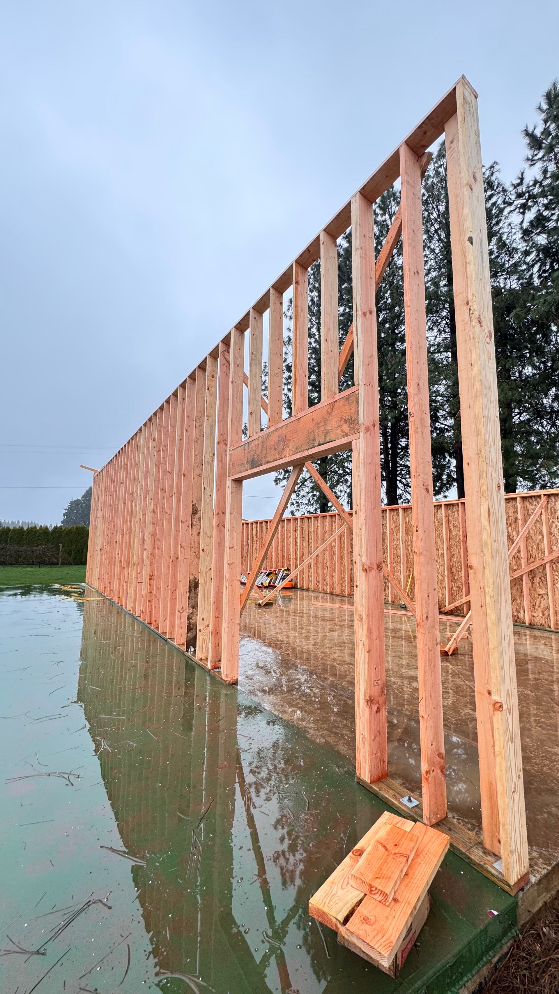 Wood-framed building under construction beside water, with exposed studs reflected on the surface.