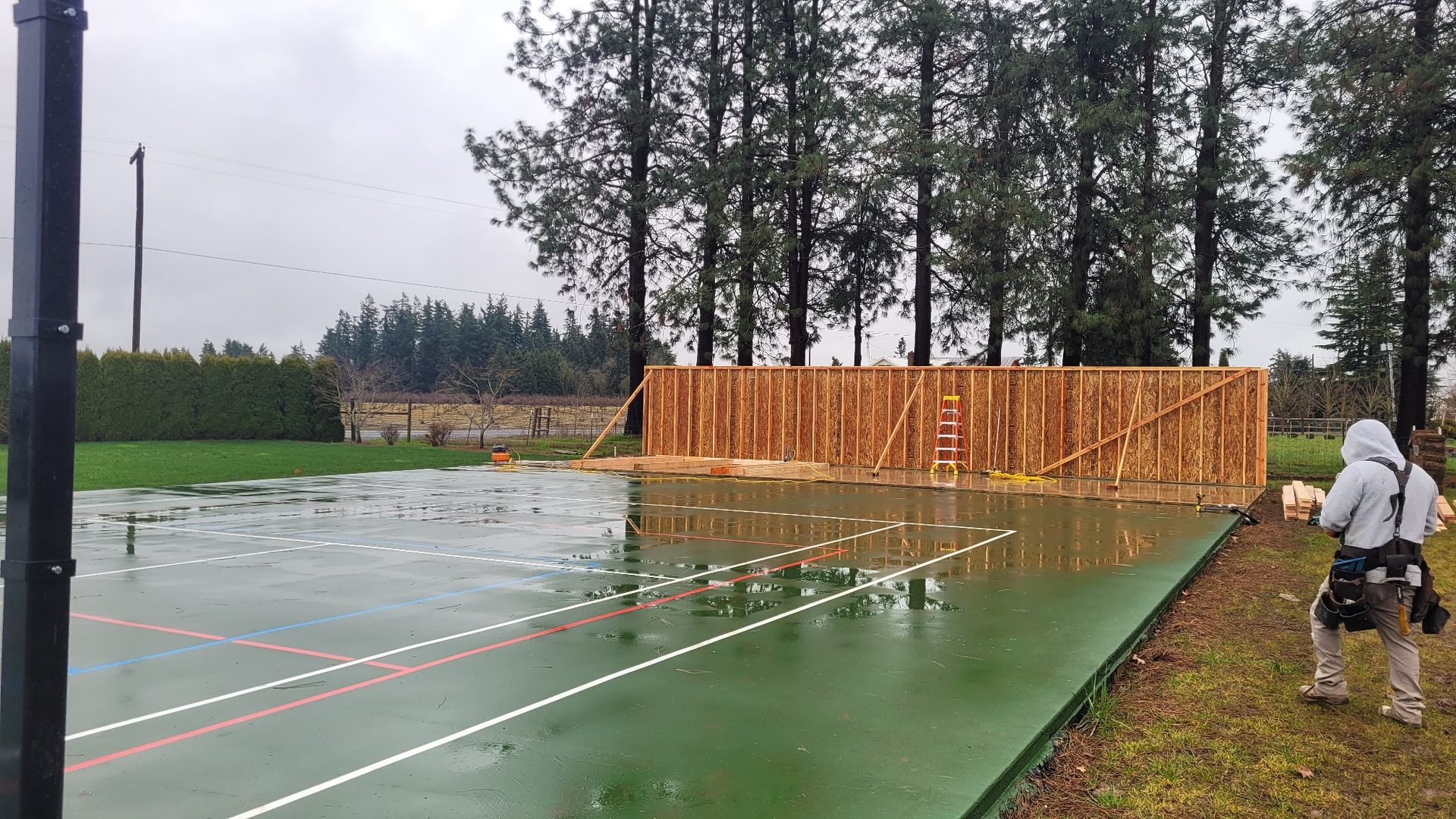 Rainy outdoor court beside an orange fence, with a person in white standing at the right edge.
