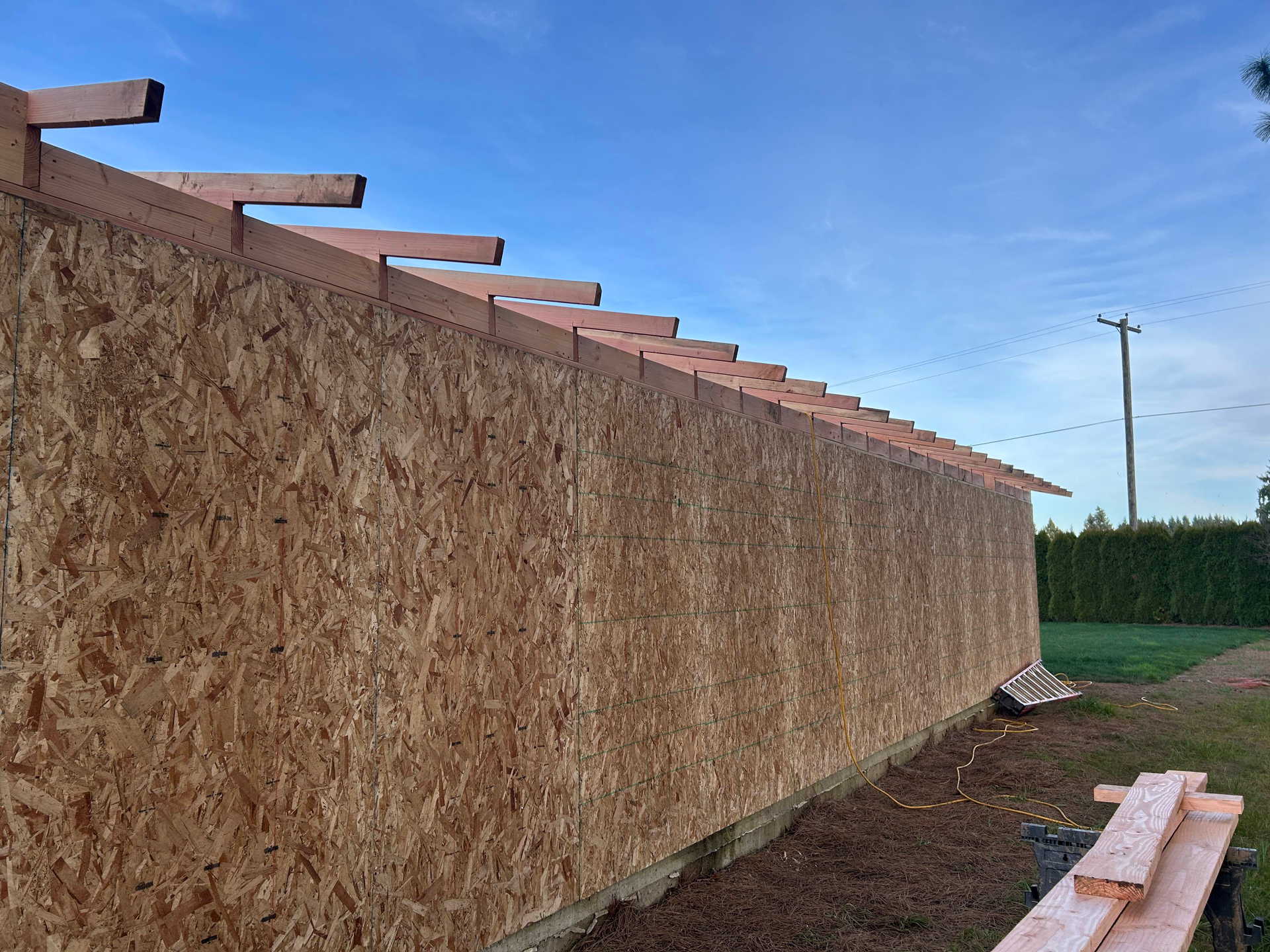 Long OSB wall under construction beside a dirt lot, with exposed roof beams and blue sky.