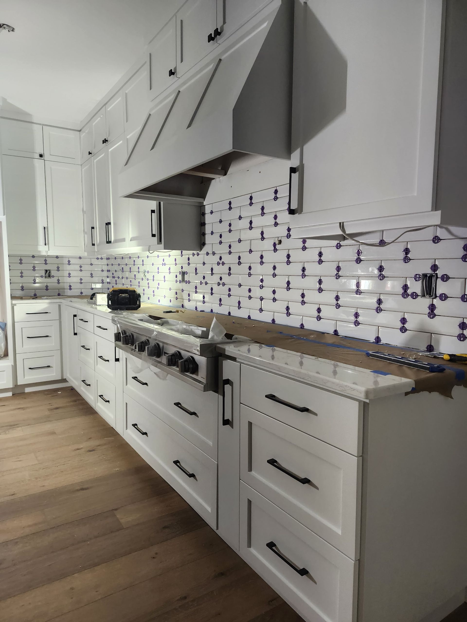 Bright white kitchen with patterned backsplash, long counter, stove, hood, and wood floor.