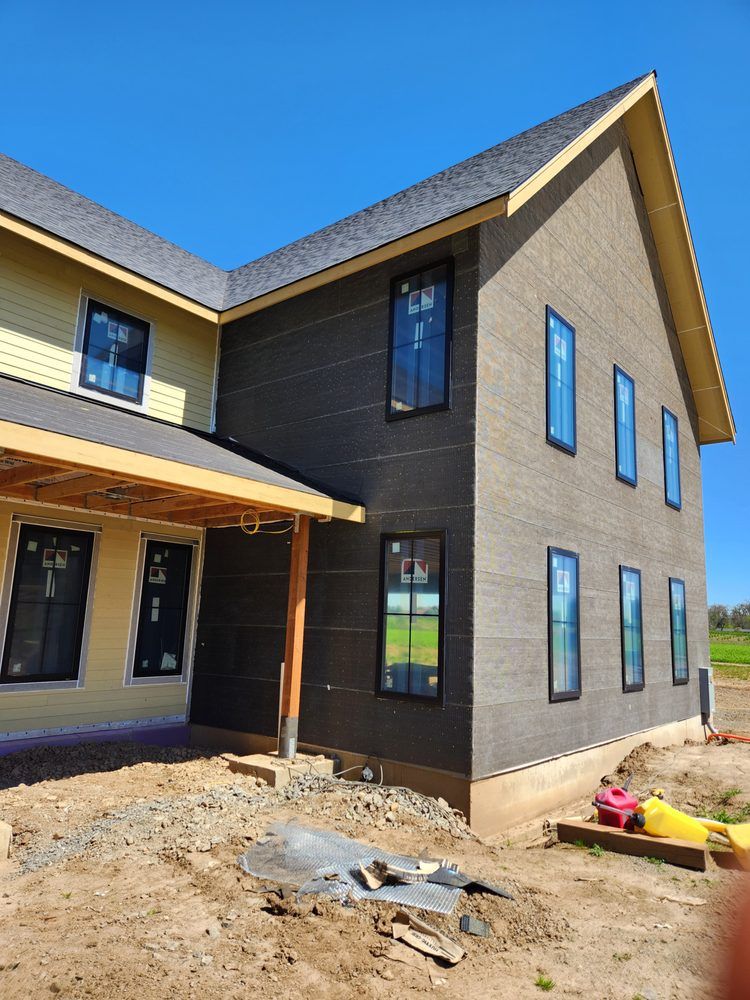 New two-story house with dark siding and tan brick exterior under construction, front porch and bare yard