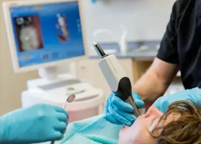 Dentist using a digital scanner in a patient's mouth; a dental assistant holds a mirror.