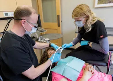 Dentist and assistant examining a patient's teeth. Dentist holds tool; assistant sits nearby. Dental office setting.