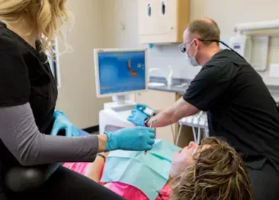 Dentist examining patient's teeth with assistant; dental office setting; computer screen shows a 3D image.