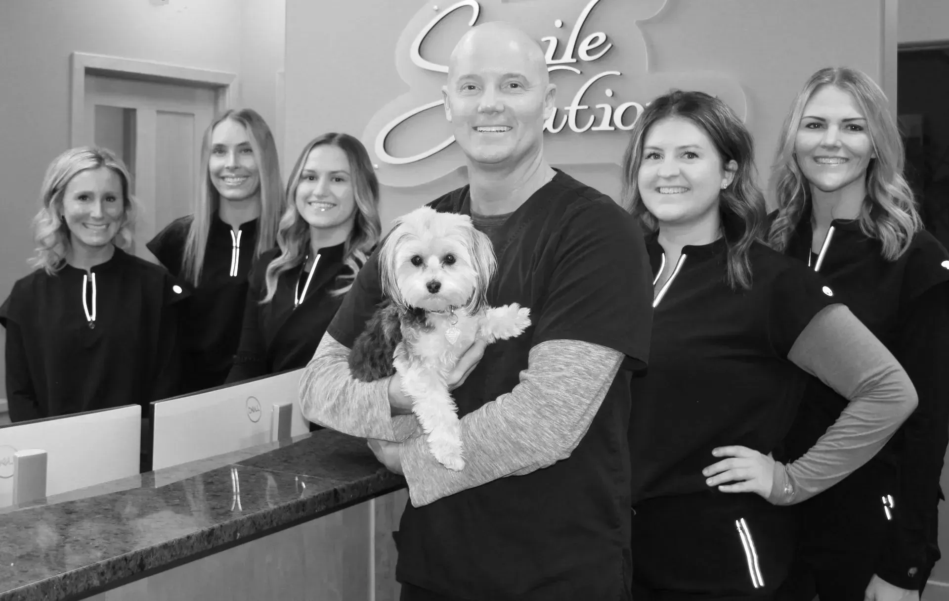 Dentist and staff with dog in office. Man holding dog smiles with 5 others in black scrubs.