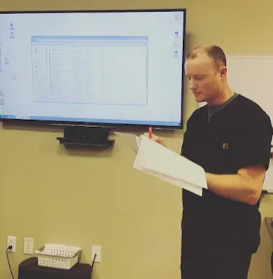 Man in black scrubs taking notes in a room with a large screen displaying a spreadsheet.