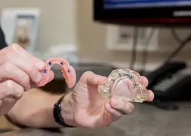 Person holding dental molds; one with pink, tooth-like structure, other clear. Office setting.