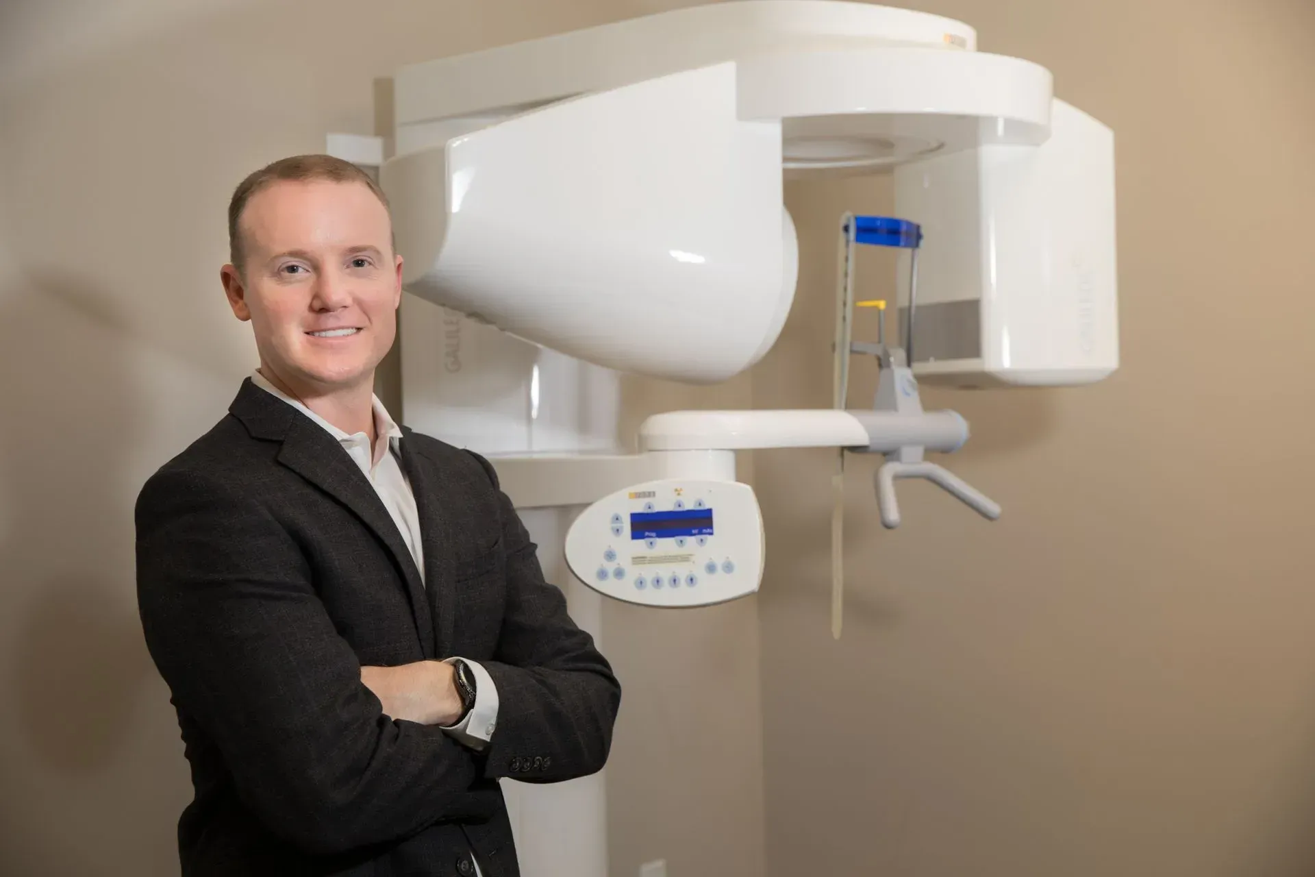 Man in suit smiles, arms crossed, stands next to a dental panoramic X-ray machine.
