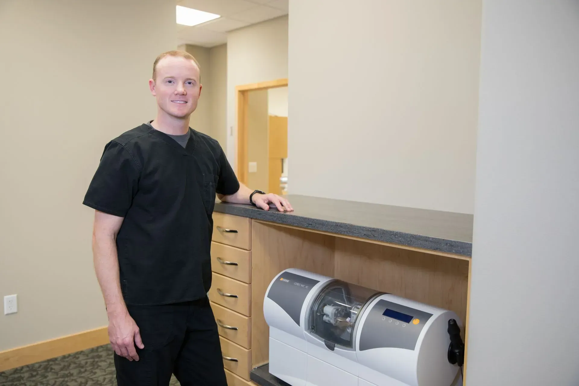 Man in black scrubs in a dental office, leaning on counter near a dental milling machine, smiling.