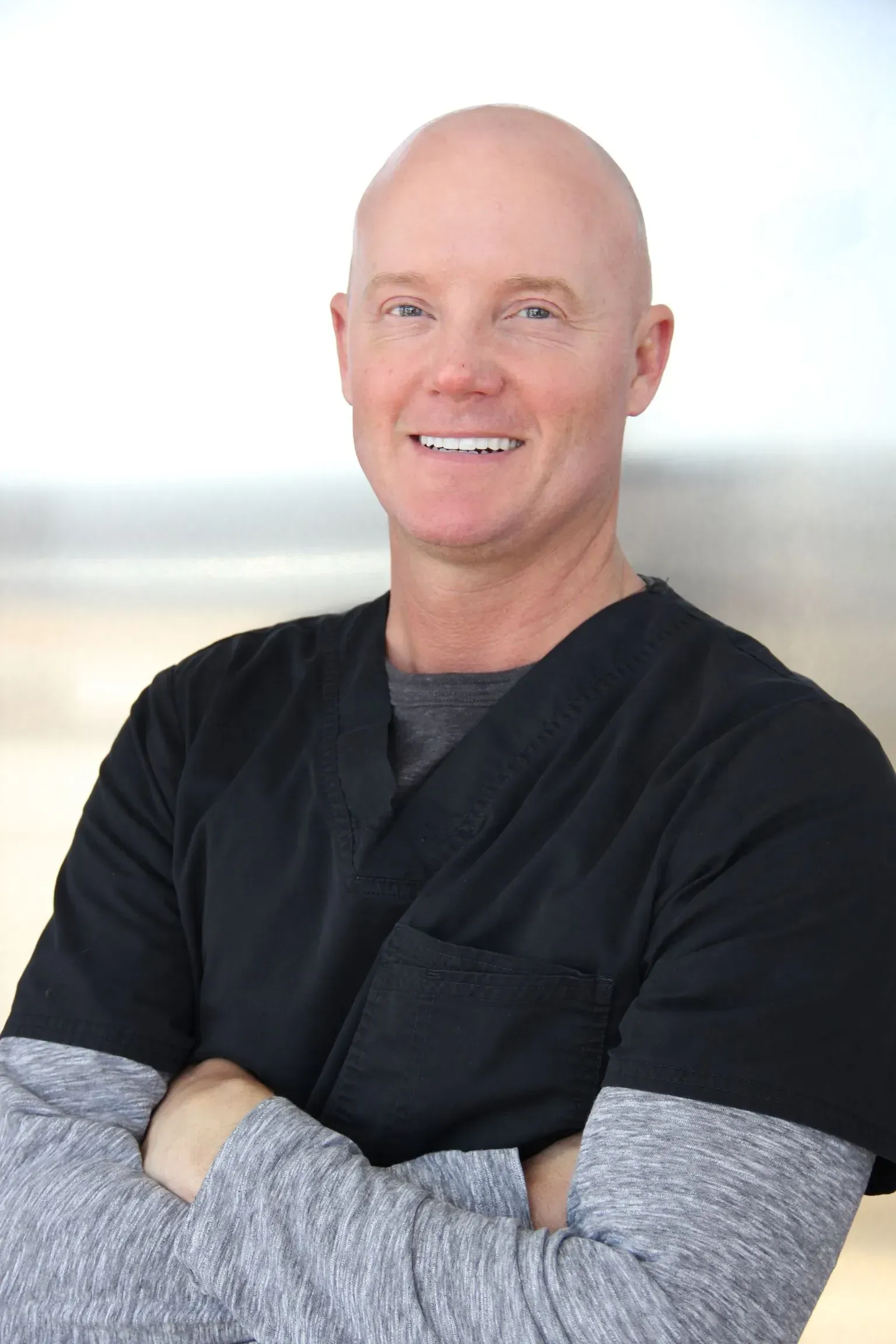 Man with crossed arms, wearing black scrubs over a grey long-sleeved shirt, smiling.