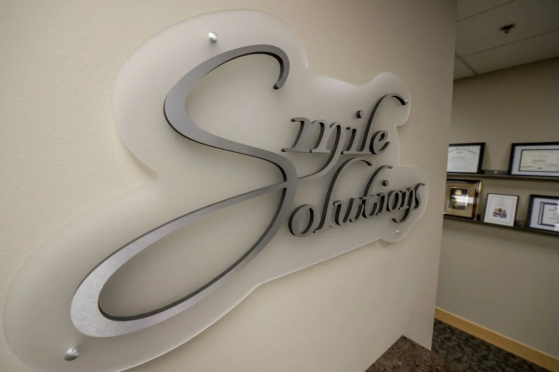 Sign for Smile Solutions dental office, silver letters and logo on a clear acrylic backing, mounted on a cream-colored wall.