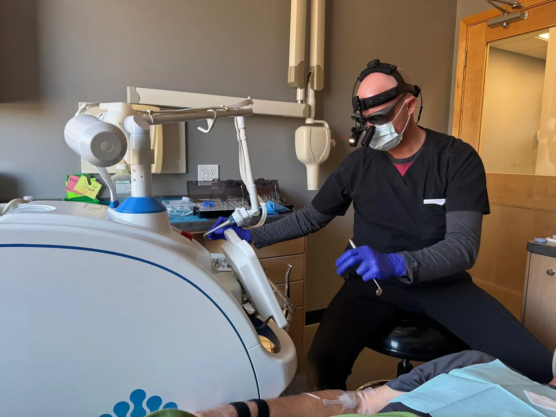 Dentist in scrubs, mask, and glasses operates dental equipment in an office setting.