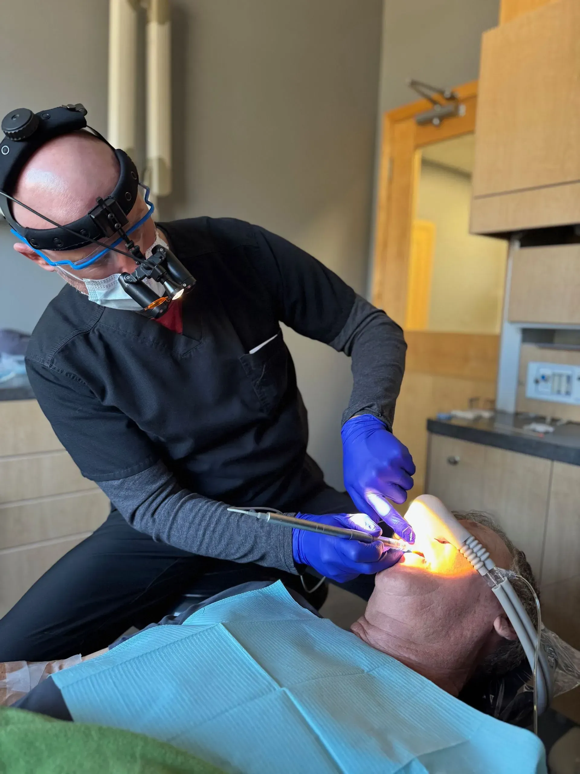 Dentist in black scrubs with headlamp, examining a patient’s mouth with tools. Blue gloves, seated.