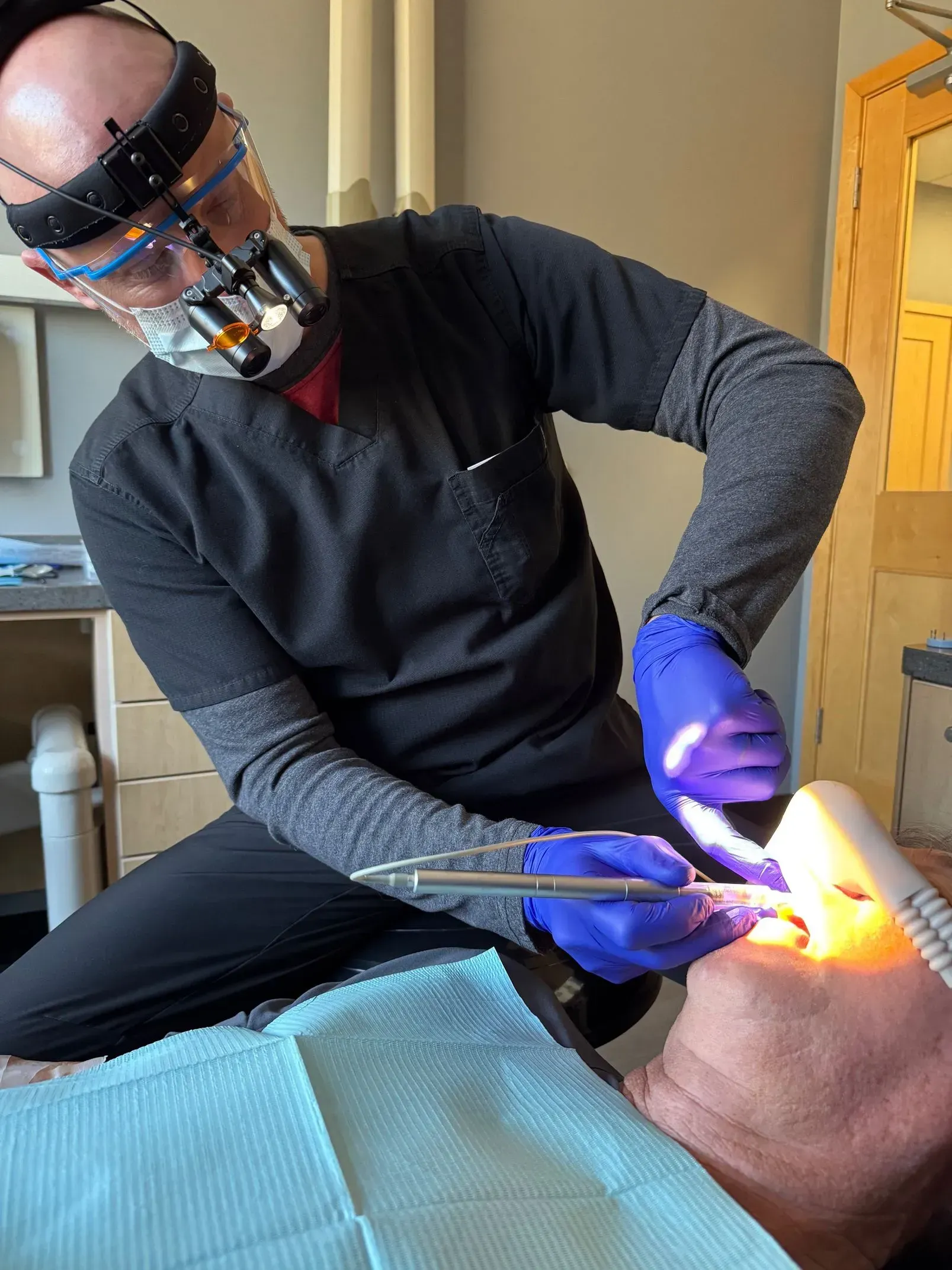 Dentist in scrubs, mask, and headlamp, working on a patient's mouth with tools. Blue gloves, dental office.