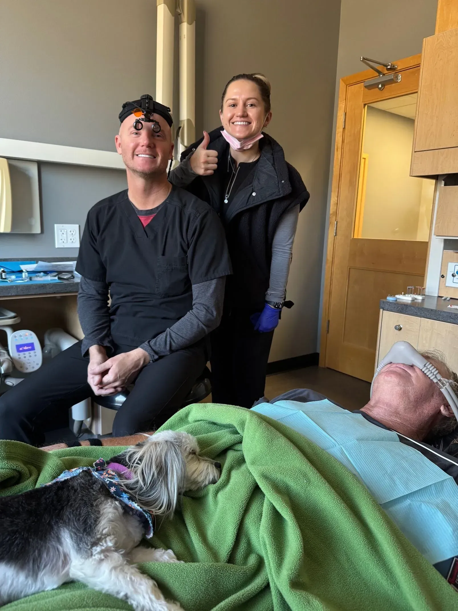 Two people in scrubs with a dog next to a patient in a dental setting.