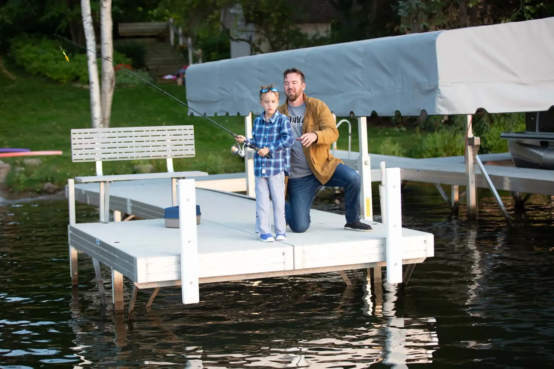 Man and child on a dock fishing. The man kneels beside the child holding a fishing rod over the water.