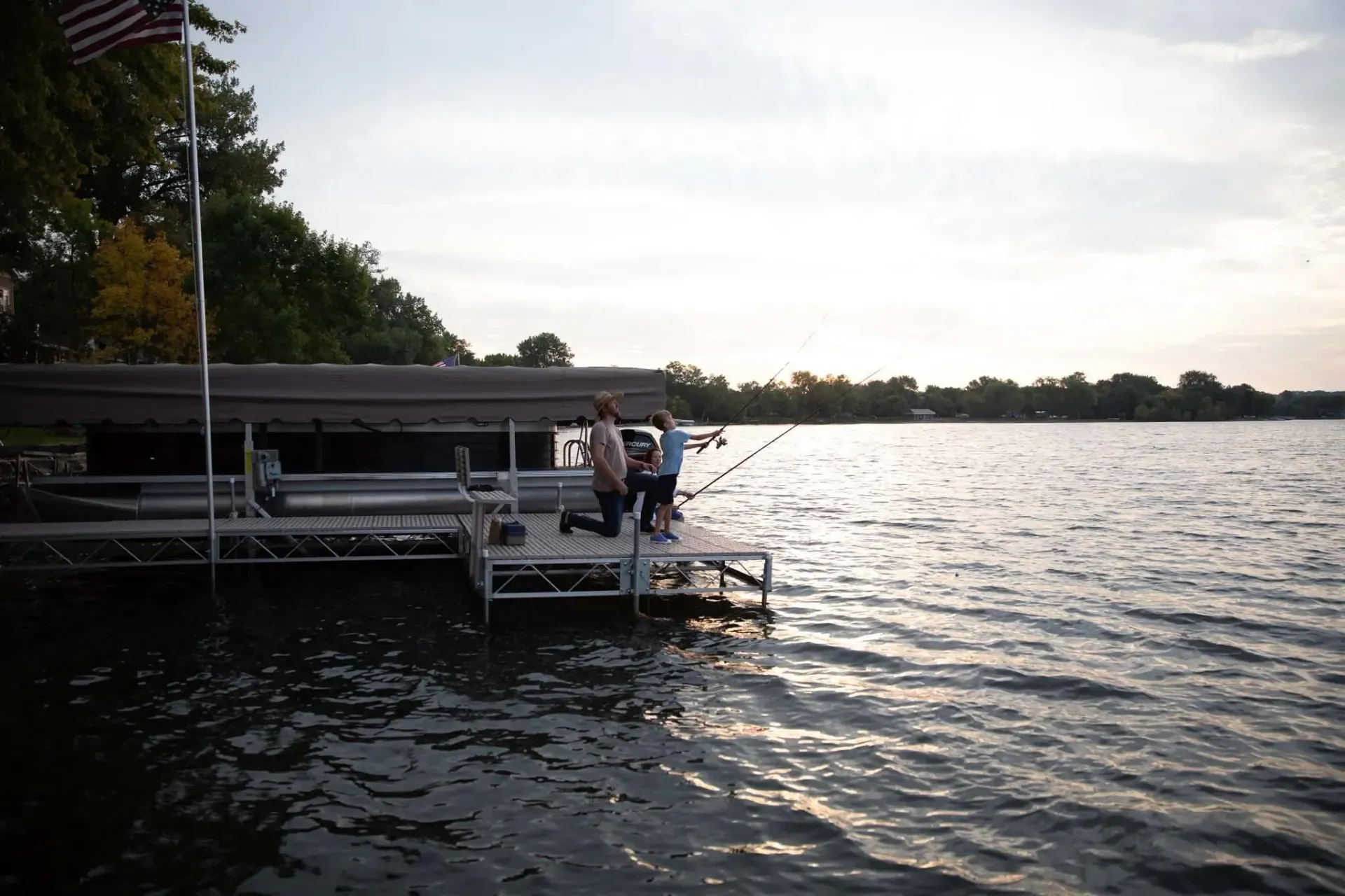 Person kneeling on a dock by a lake, gesturing. Sunset casts a warm glow; ripples on the water.