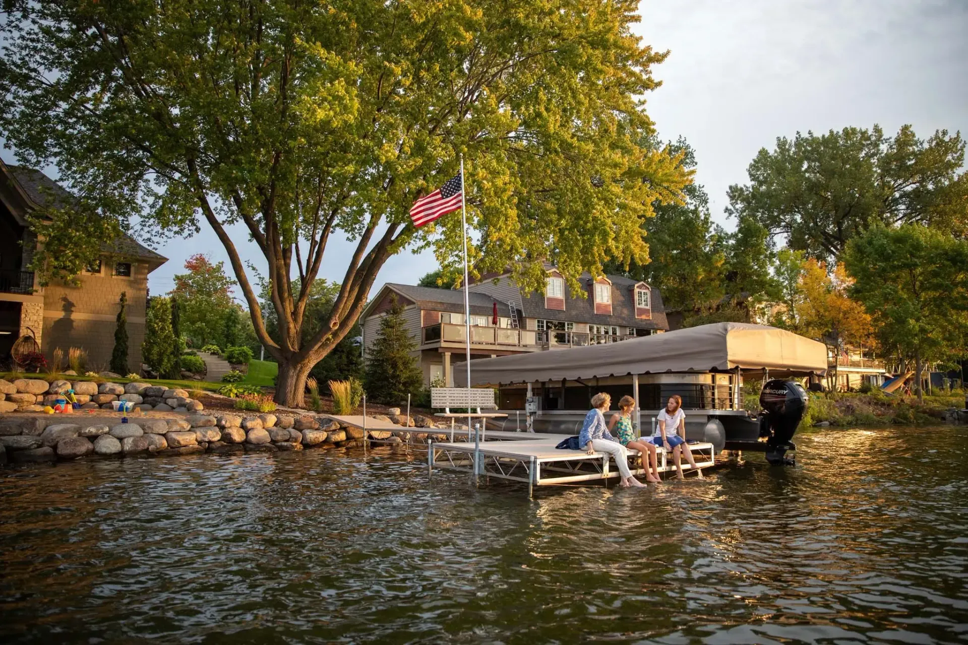 People sitting on a dock by a lake, beneath a covered boat lift with a house and trees in the background.