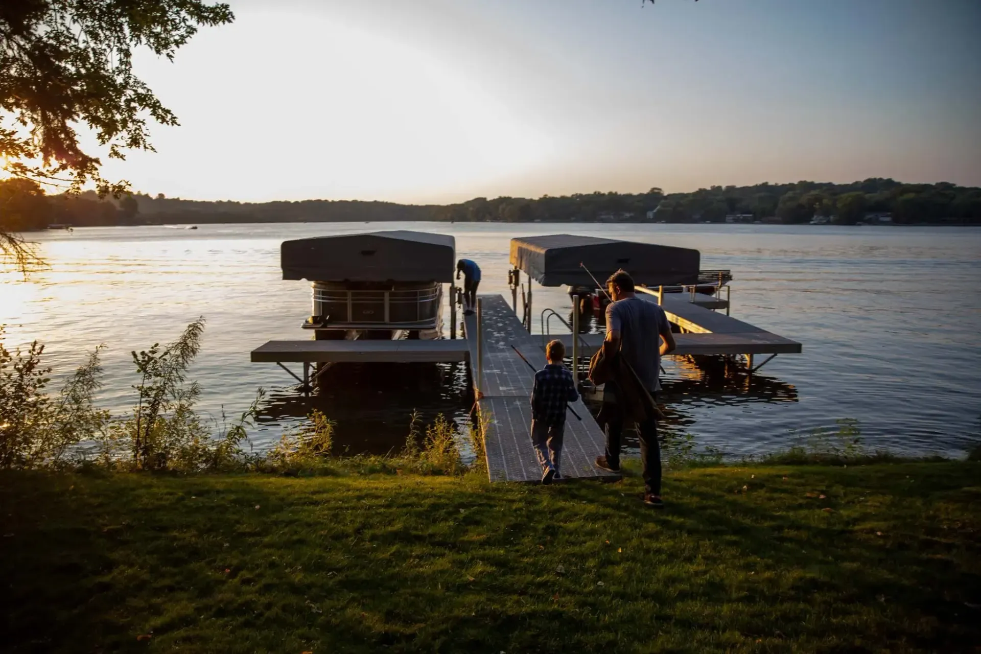 People walking towards a dock with boat lifts on a lake at sunset.