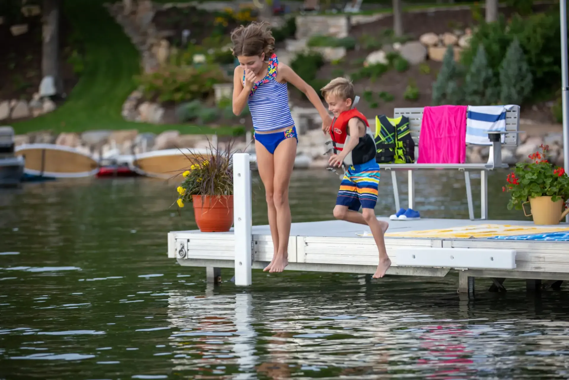Two children jump into a lake from a dock. The girl wears a blue swimsuit, boy wears swim trunks and a life vest.