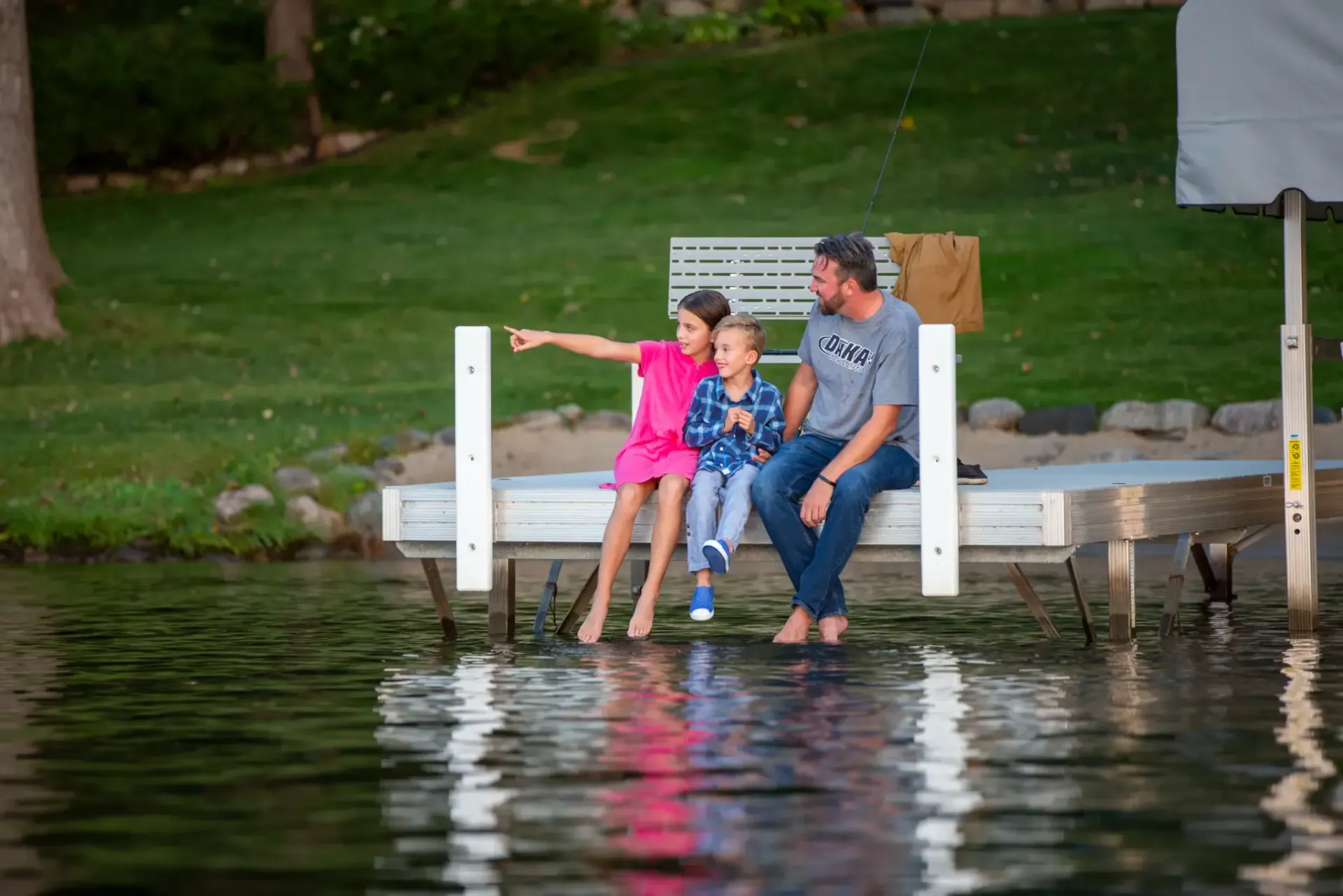 Family of three sits on a dock, feet in the water, looking at something off-screen.