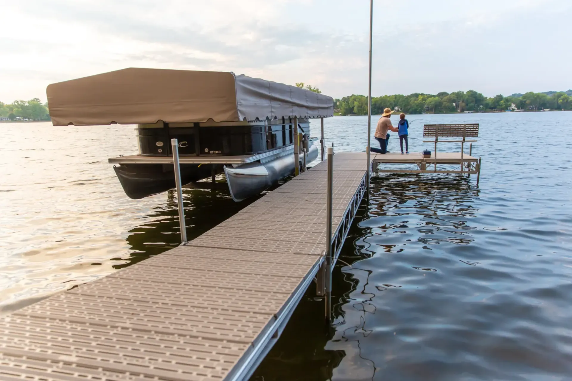 Pontoon boat on lift at dock, two people near end of dock on water, overcast sky.