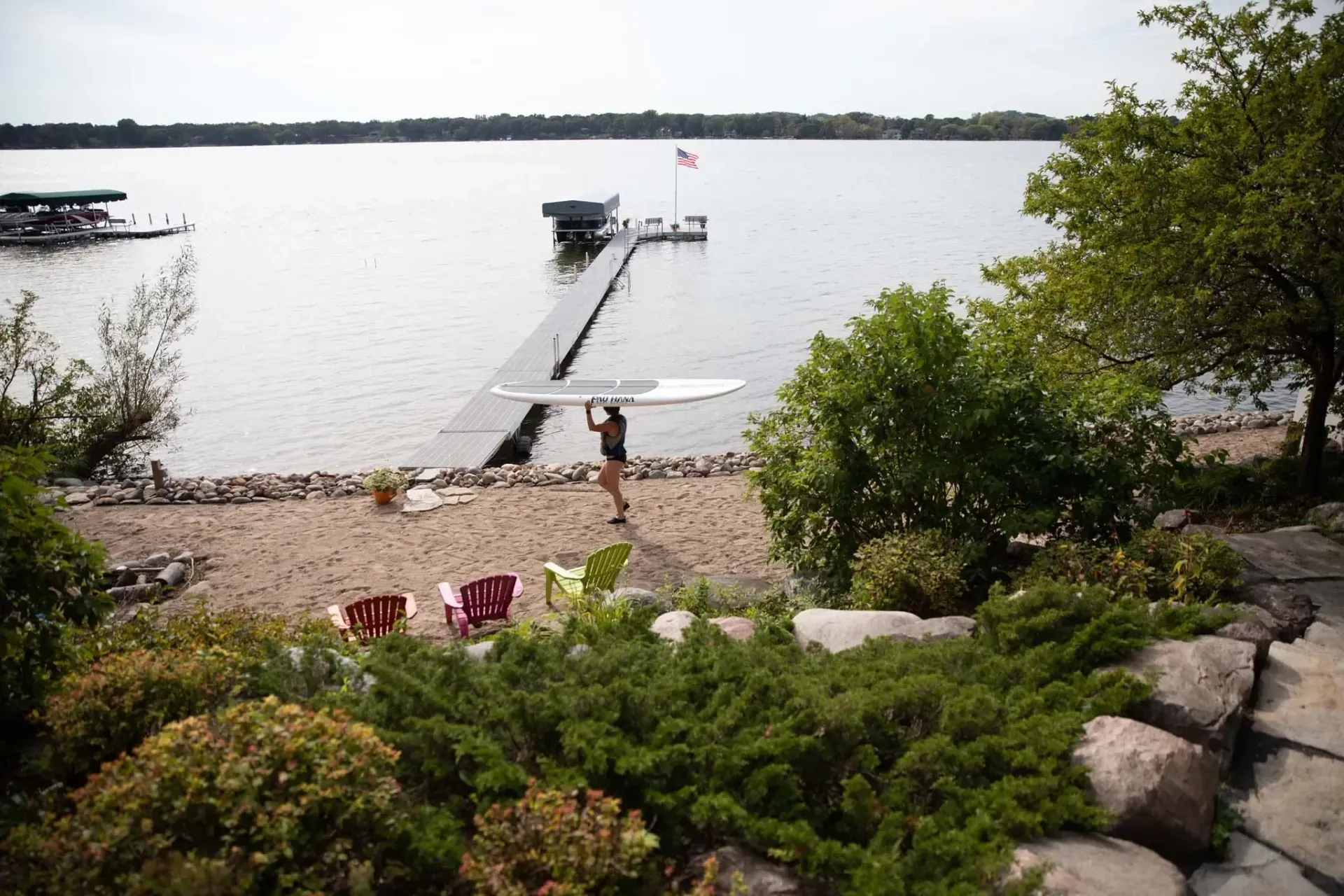 Man carrying large white object near lake and dock, with shore and vegetation in foreground.