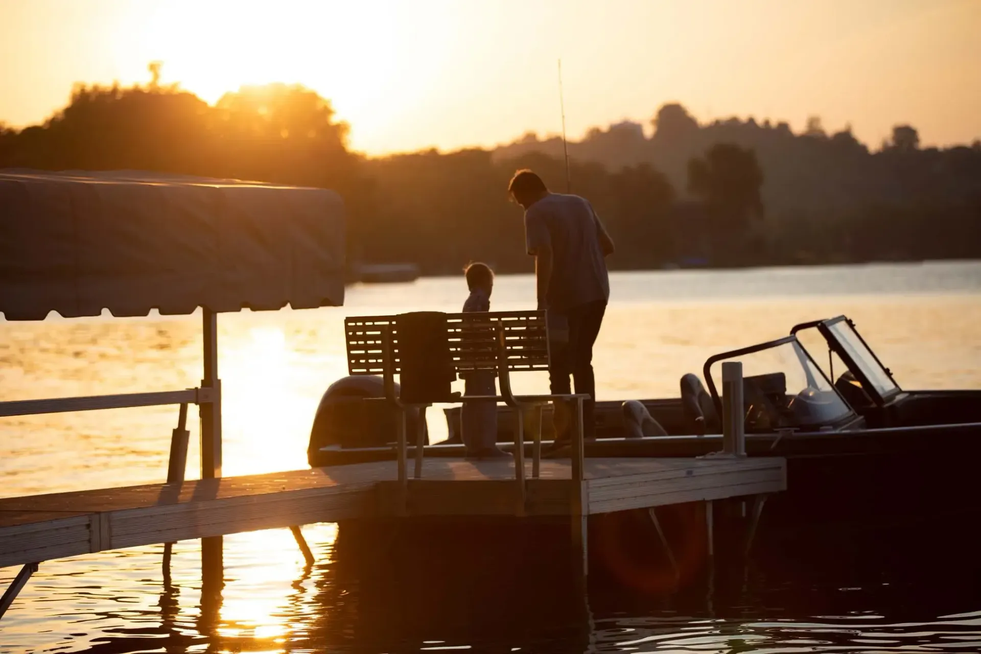Sunset over a lake with two figures on a dock, one appearing to be leaning or observing something.