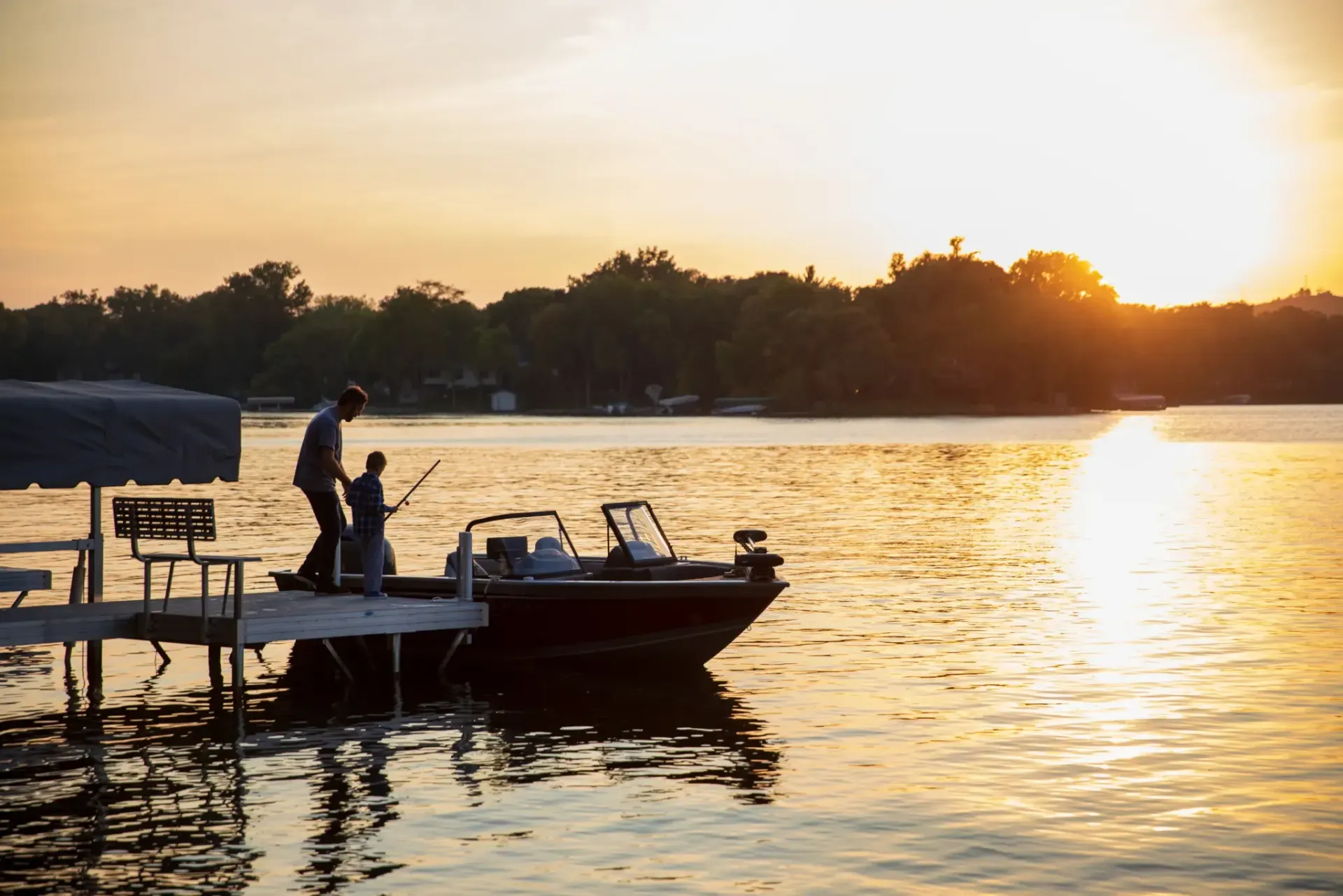 Silhouette of two people fishing from a dock at sunset, next to a boat on calm water.
