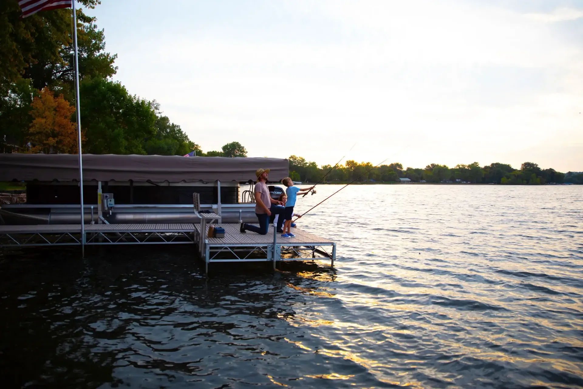 People on a dock near a lake at sunset. A boat is stored on the dock, flag.