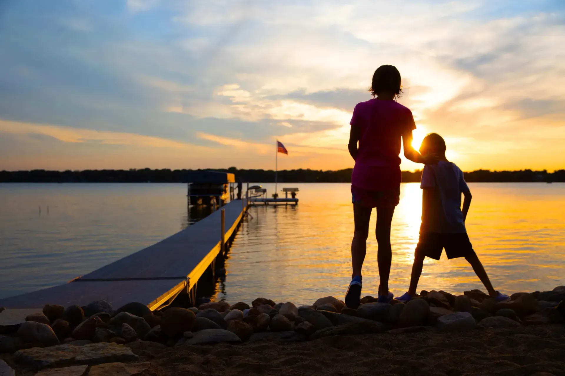 Silhouette of two people by a lake at sunset. A pier extends into the water.