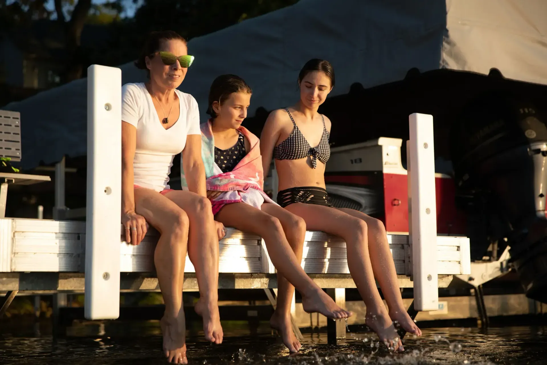 Three people sitting on a dock with their feet in the water; a woman, girl, and another woman.