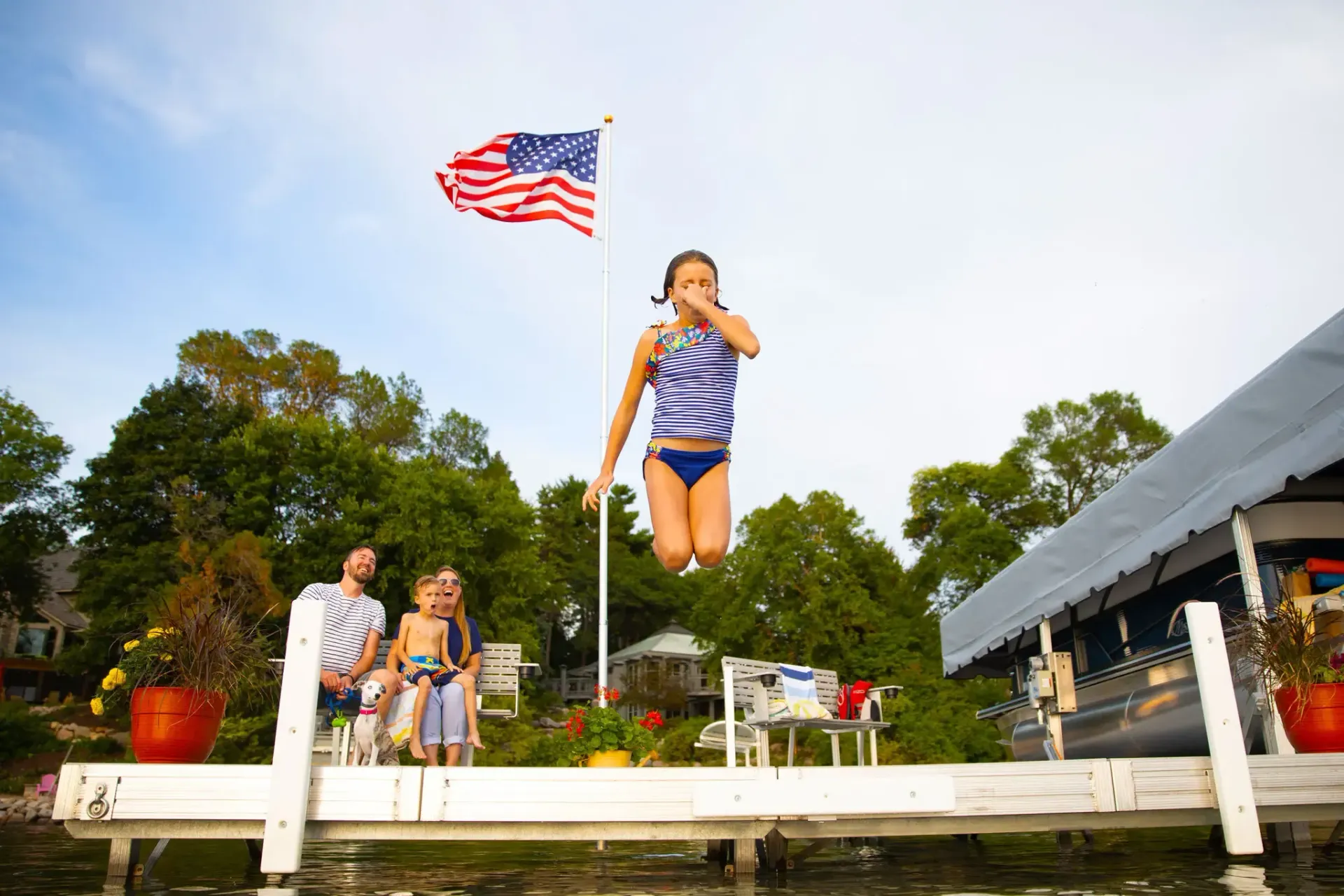 Girl jumping off a dock into water, American flag in background, family watching.