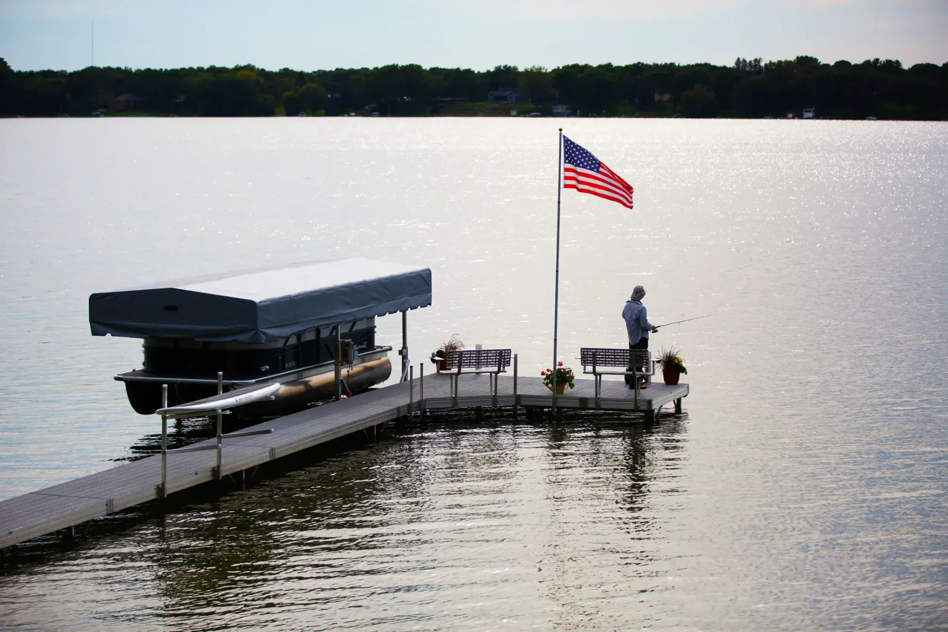 Dock with boat lift, American flag, and person fishing on a lake under a bright sky.