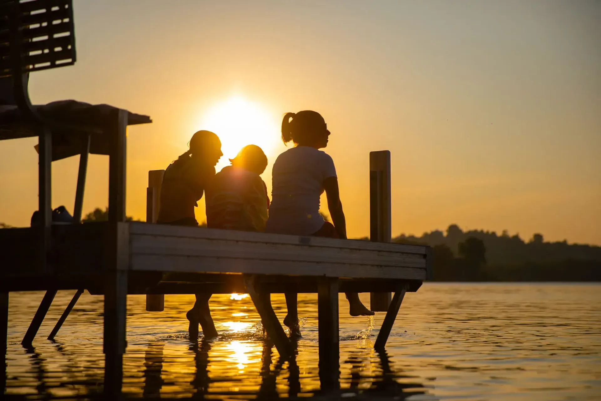 Three people silhouetted on a wooden dock at sunset, overlooking calm water.